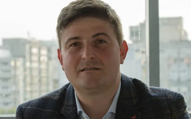 Un bărbat cu părul scurt și sacou închis peste o cămașă deschisă stă în fața unei ferestre cu clădiri în fundal.