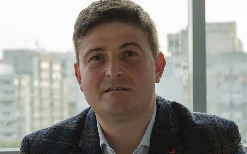 Un bărbat cu părul scurt și sacou închis peste o cămașă deschisă stă în fața unei ferestre cu clădiri în fundal.