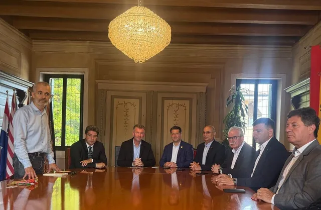Opt bărbați stau la o masă lungă de lemn într-o încăpere cu un candelabru suspendat și ferestre mari în fundal.