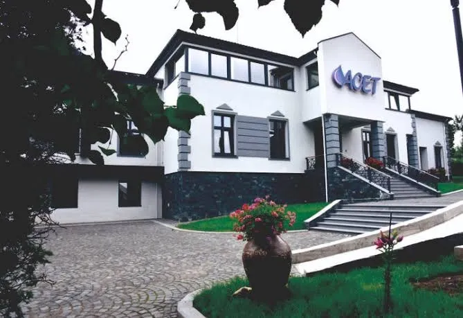 O clădire albă cu ferestre negre, un logo albastru pe fațadă și scări exterioare cu balustrade metalice, înconjurat de vegetație și un pavaj de piatră.