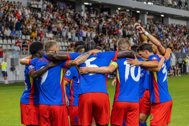 Un grup de fotbaliști în echipament sportiv albastru și roșu stau cu spatele, îmbrățișându-se pe un teren de fotbal în fața unui stadion aglomerat.