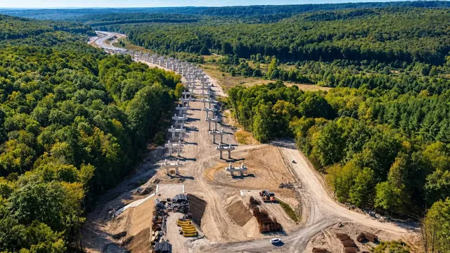 O vedere aeriană prezintă o secțiune de autostradă în construcție cu piloni de beton și drumuri de acces prin pădure.