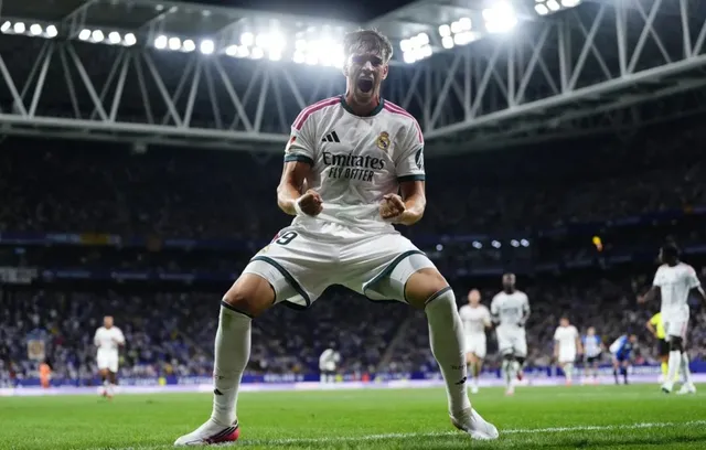 Un fotbalist într-un echipament alb de joc sărbătorește pe un teren de fotbal sub luminile unui stadion plin de spectatori.