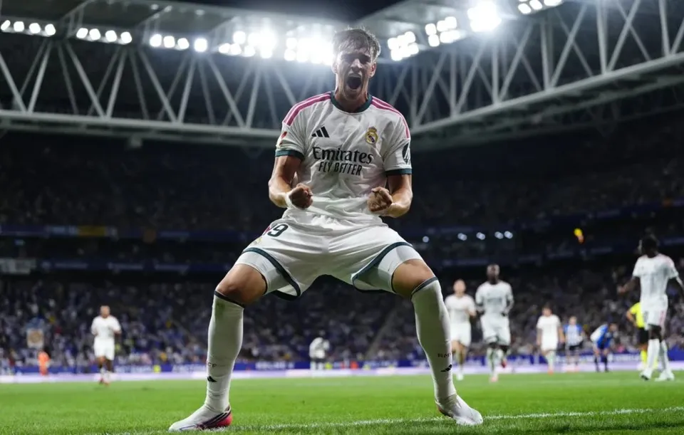 Un fotbalist într-un echipament alb de joc sărbătorește pe un teren de fotbal sub luminile unui stadion plin de spectatori.