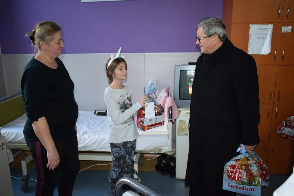 O femeie, o fată și un bărbat stau într-o cameră lângă un pat cu lenjerie albă, purtând haine casual și un sac cu produse alimentare.