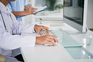 O persoană în halat alb de medic folosește un mouse și un tastatură pe un birou lângă un monitor de calculator.