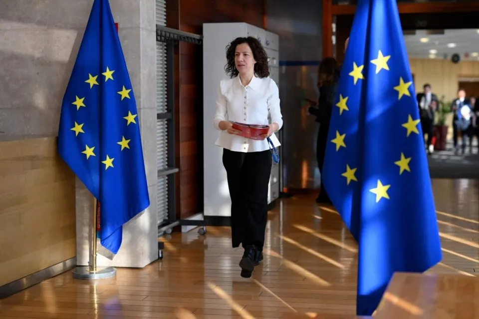 O femeie cu părul șaten, purtând o cămașă albă și pantaloni negri, merge pe un hol cu podea de lemn între două steaguri ale Uniunii Europene.