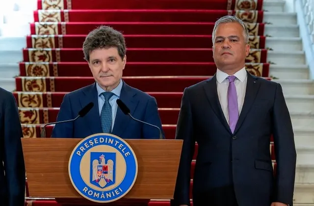 Un bărbat cu părul scurt și gri stă în picioare lângă un alt bărbat cu părul brun, amândoi purtând costume și cravate.