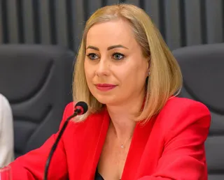 O femeie cu părul blond și sacou roșu stă la o masă lângă un microfon negru.