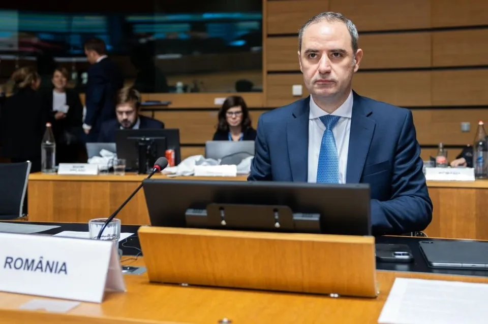 Un bărbat într-un costum albastru și cravată albastră stă la o masă de conferință lângă un monitor și un semn cu textul ROMÂNIA.