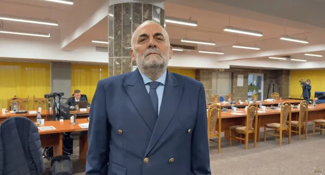 Un bărbat cu barbă și costum albastru stă în fața unor mese de conferință într-o încăpere iluminată de corpuri de iluminat lungi.