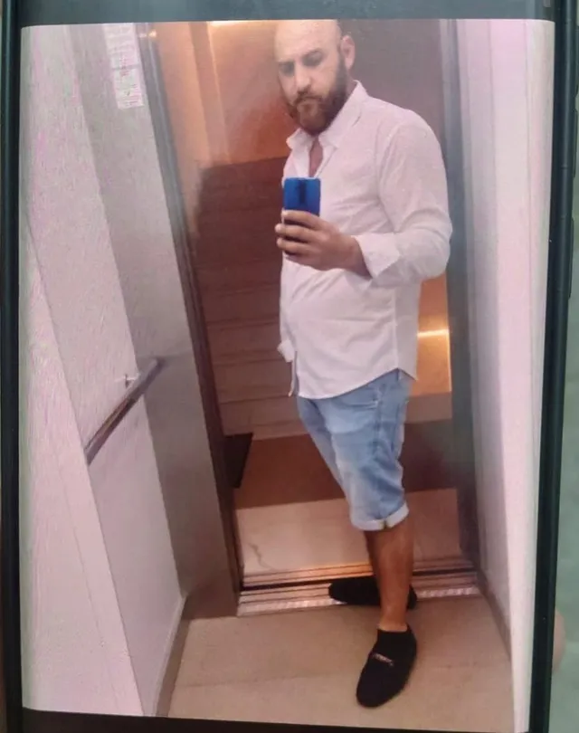 Un bărbat cu barbă, purtând o cămașă albă și pantaloni scurți albaștri, își face o fotografie în oglindă cu un telefon albastru.