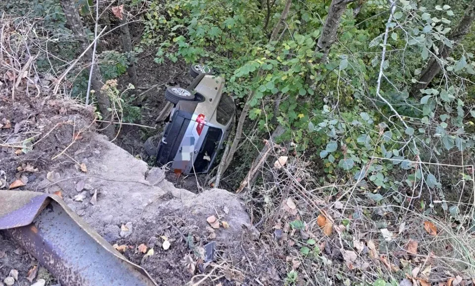 Un autoturism de culoare închisă este răsturnat pe o pante împădurită, situat sub marginea unui drum de țară.