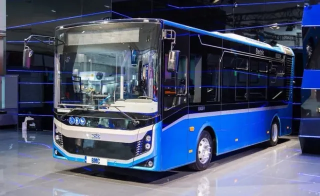 Un autobuz albastru și alb cu inscripția Electric este parcat într-un spațiu interior cu podea lucioasă.