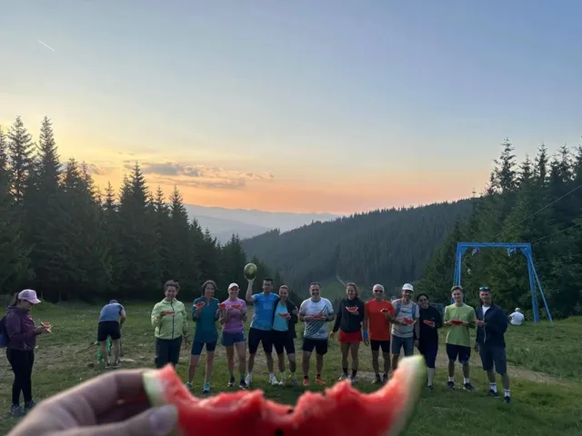 Un grup de oameni stă în linie pe o prăpastie verde sub un cer la apus, în timp ce o felie de pepene este ținută în prim-plan.