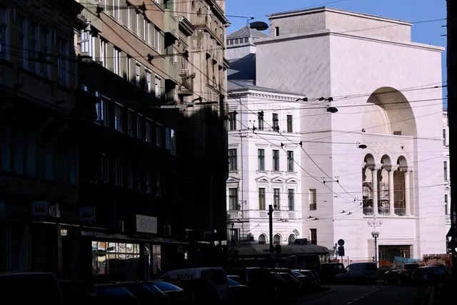 O clădire albă cu un arc mare domină fundalul, fiind încadrată de alte edificii și de cabluri suspendate deasupra unei străzi cu mașini parcate.