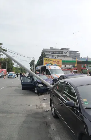 Un autoturism negru este oprit lângă o ambulanță albă sub un stâlp înclinat pe o stradă cu clădiri în fundal.