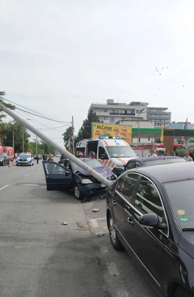 Un autoturism negru este oprit lângă o ambulanță albă sub un stâlp înclinat pe o stradă cu clădiri în fundal.