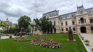 O clădire monumentală de culoare deschisă este situată în spatele unui gazon verde cu flori aranjate în forme curbate și un bust pe un soclu.