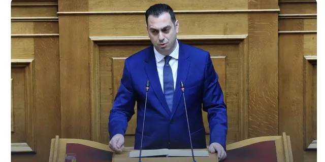 Un bărbat cu sacou albastru și cravată stă în spatele unui pupitru în fața unui perete din panouri de lemn.