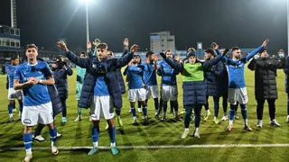 Un grup de fotbaliști în echipament sportiv albastru și alb stau pe un teren de fotbal sub luminile stadionului cu brațele ridicate.