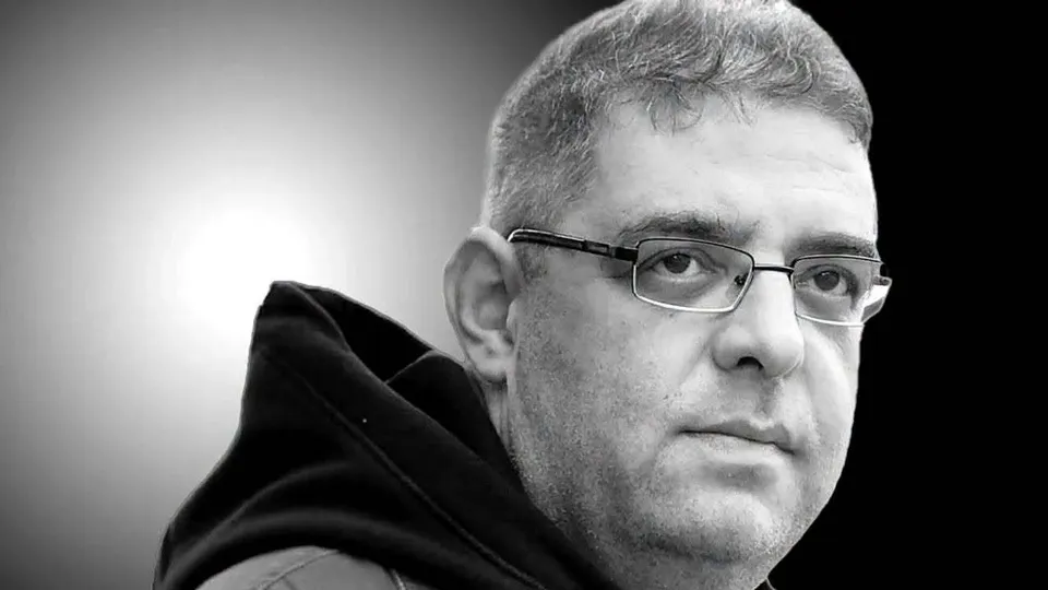 Un bărbat cu ochelari și păr scurt, purtând o haină închisă la culoare, este surprins într-un prim-plan de tip portret pe un fundal gri.