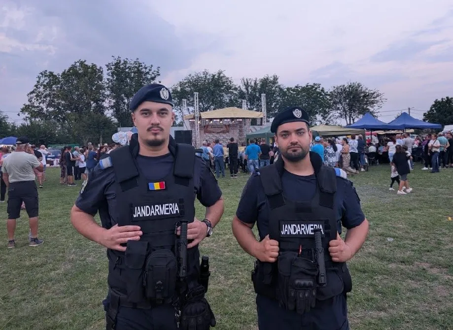 Doi bărbați în uniformă neagră cu veste tactice și berete stau în picioare pe un teren de iarbă în fața unei mulțimi.