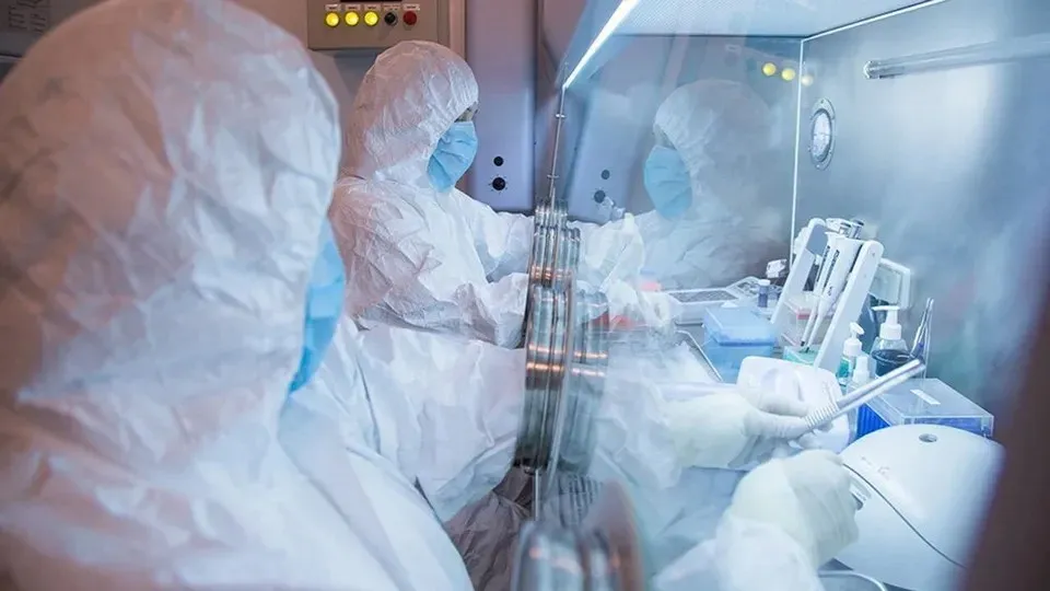 Două persoane purtând costume de protecție albe, măști și mănuși de protecție lucrează la un set de echipamente medicale într-un mediu steril.
