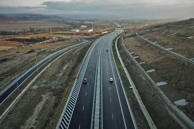 O vedere aeriană prezintă o autostradă cu două benzi pe fiecare sens, traversând un peisaj de dealuri cu vegetație uscată și câteva vehicule circulând.