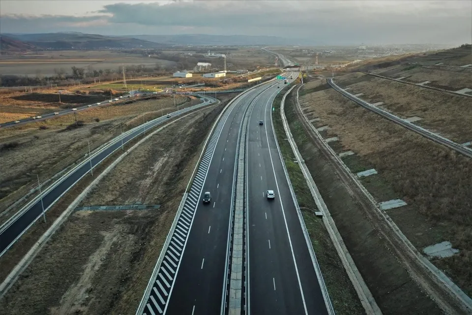 O vedere aeriană prezintă o autostradă cu două benzi pe fiecare sens, traversând un peisaj de dealuri cu vegetație uscată și câteva vehicule circulând.