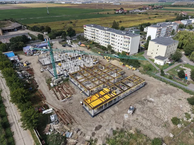 O vedere aeriană prezintă un șantier cu structuri de beton și cadre metalice de culoare galbenă lângă câteva blocuri de locuințe.
