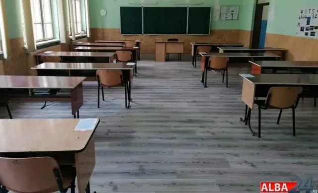 O sală de clasă cu mai multe birouri de lemn și scaune maronii dispuse pe un podea de lemn, având un panou verde pe perete.