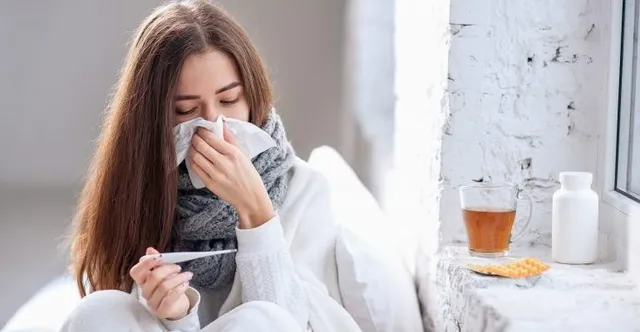 O femeie cu părul lung, purtând un șal gri și haine albe, ține un termometru și șterge nasul cu un șervețel de hârtie lângă o cană.