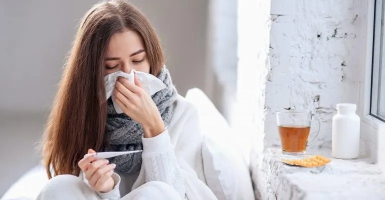 O femeie cu părul lung, purtând un șal gri și haine albe, ține un termometru și șterge nasul cu un șervețel de hârtie lângă o cană.