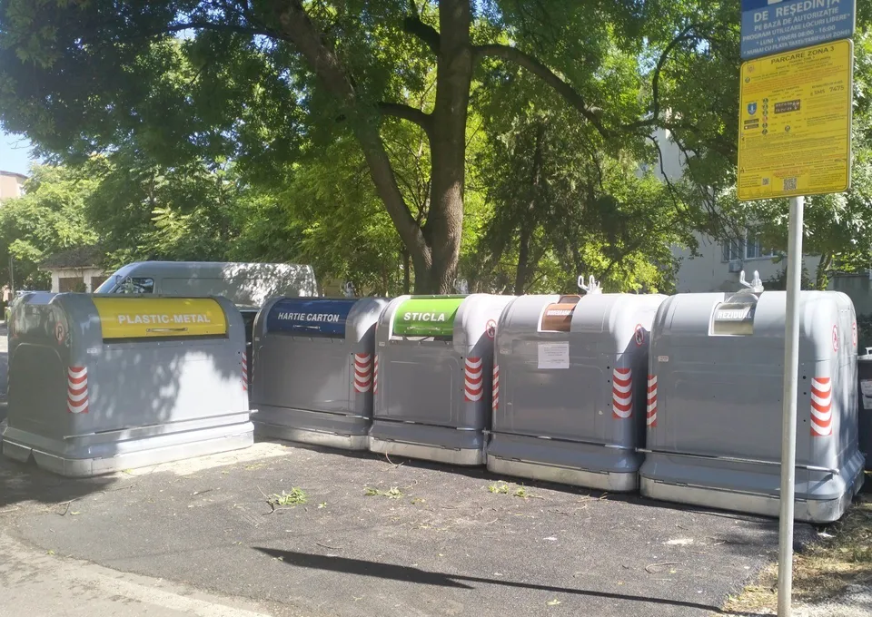 Cinci containere de reciclare gri cu etichete colorate sunt aliniate pe un teren asfaltat sub copaci.