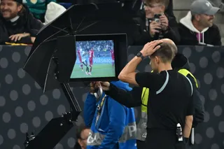 Un bărbat cu tricou negru stă în spatele unui monitor care afișează o scenă de pe un teren de fotbal sub o umbrelă neagră.