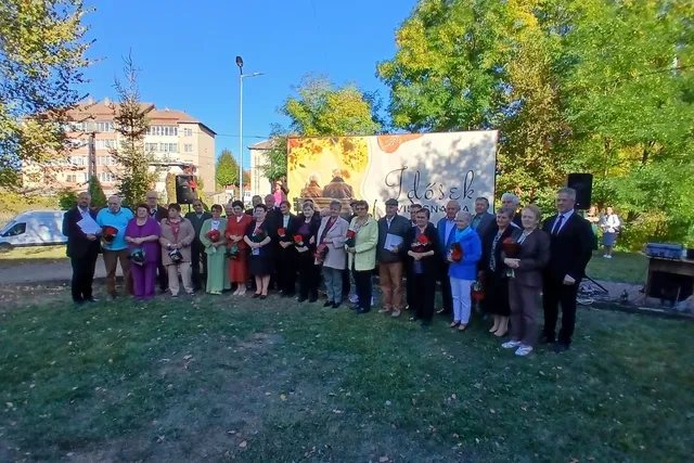 Un grup de persoane stă în linie pe un teren cu iarbă în fața unui panou decorativ mare și a unor copaci verzi.