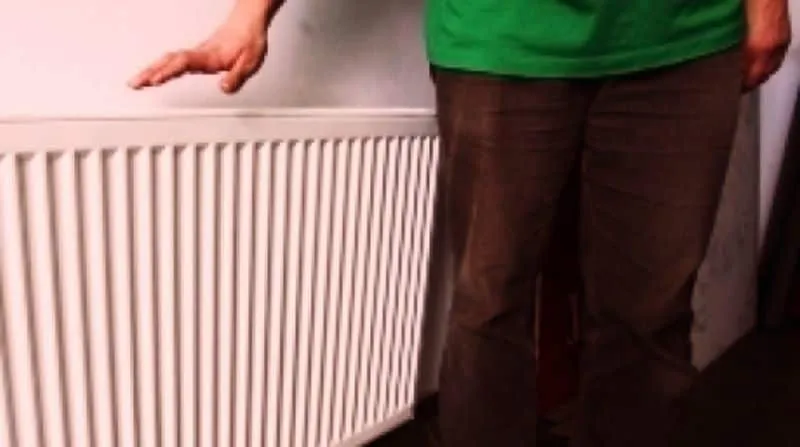 O persoană purtând un tricou verde și pantaloni maro stă lângă un calorifer alb de perete.