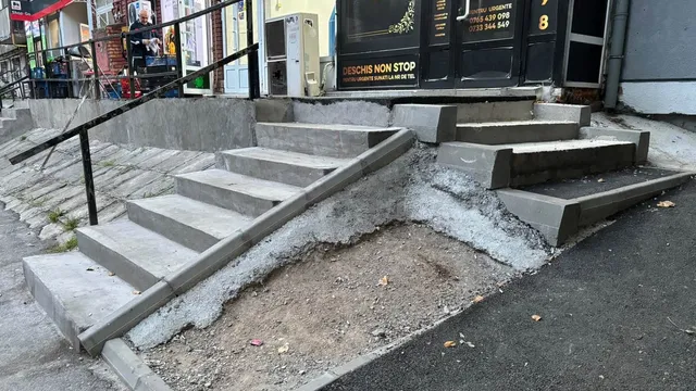 Scări de beton cu trepte de dimensiuni diferite sunt construite pe un teren neuniform lângă un trotuar și o clădire.
