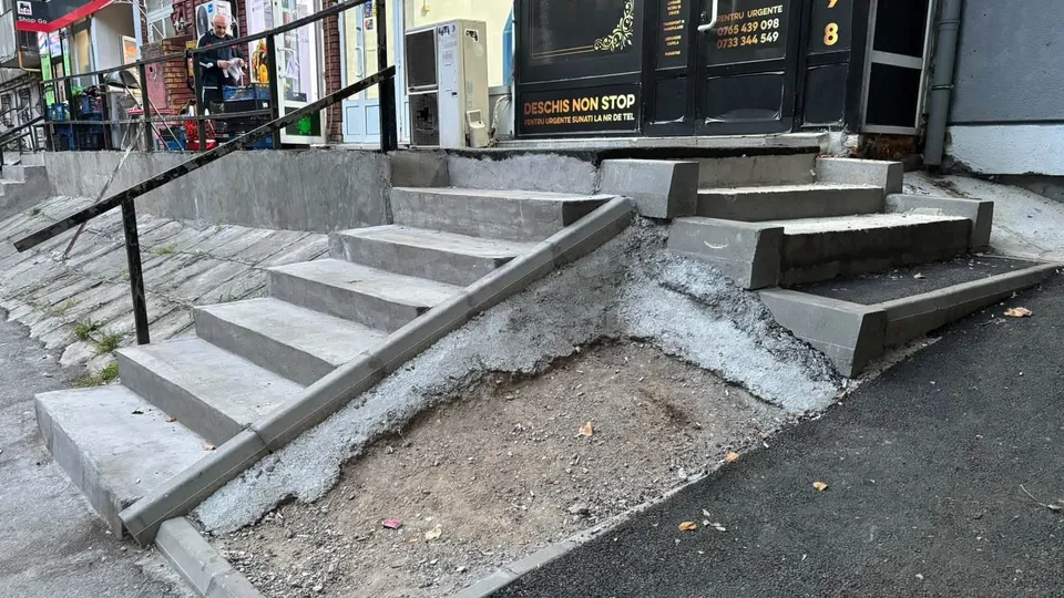 Scări de beton cu trepte de dimensiuni diferite sunt construite pe un teren neuniform lângă un trotuar și o clădire.