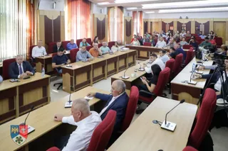 O sală de conferințe plină cu oameni așezați la mese lungi, cu microfoane individuale, indicând o întâlnire sau o ședință.