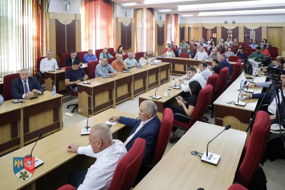 O sală de conferințe plină cu oameni așezați la mese lungi, cu microfoane individuale, indicând o întâlnire sau o ședință.