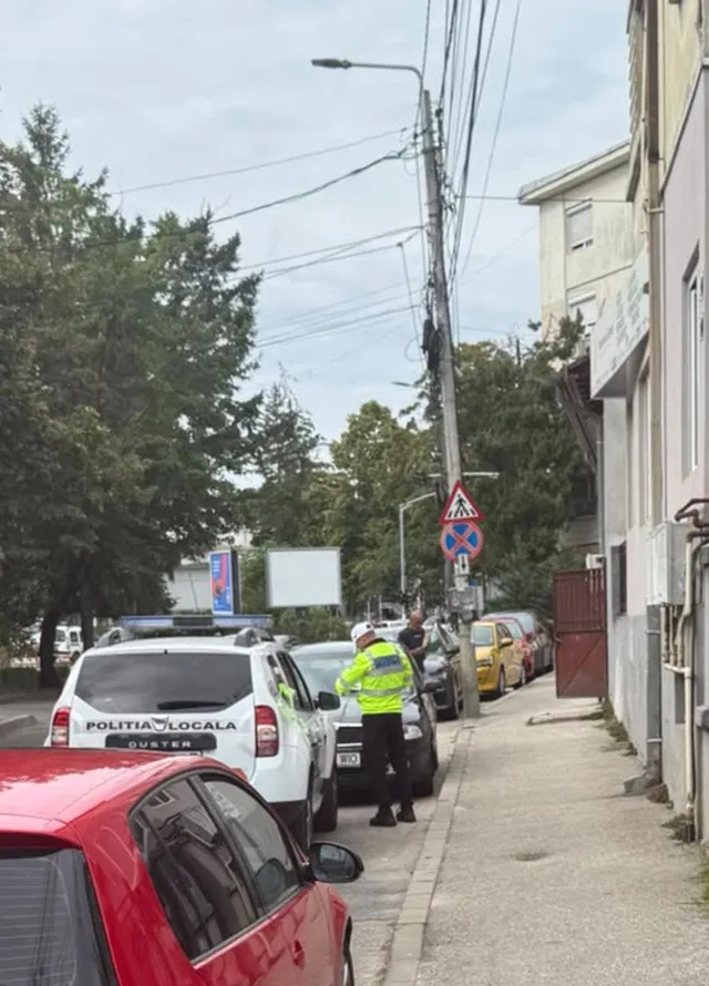 Un agent în uniformă galbenă stă pe trotuar lângă mașini parcate într-o zonă cu clădiri și copaci.