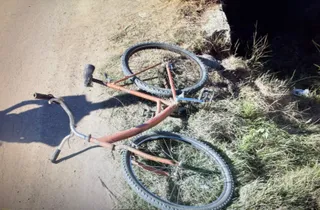 O bicicletă de culoare portocalie cu roți negre este întinsă pe un drum de pământ lângă o zonă cu iarbă uscată și un taluz.