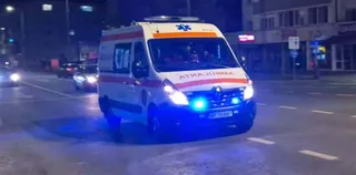 O ambulanță albă cu lumini albastre aprinse circulă pe un drum asfaltat noaptea, lângă alte vehicule în fundal.
