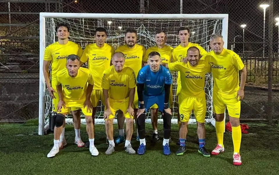 O echipă de nouă bărbați îmbrăcați în echipamente sportive galbene și unul în albastru pozează în fața unei porți de fotbal pe un teren sintetic.