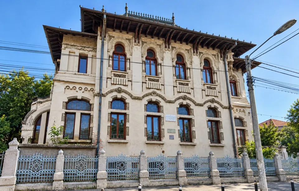 O clădire istorică de două etaje cu arhitectură neogotică, ferestre cu arcade decorative, acoperiș înalt și un gard de metal forțat cu stâlpi de piatră.