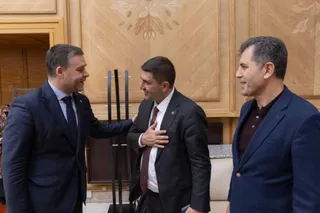 Trei bărbați în costume purtând cămași și cravate stau în picioare într-o încăpere cu panouri de lemn decorat pe fundal.