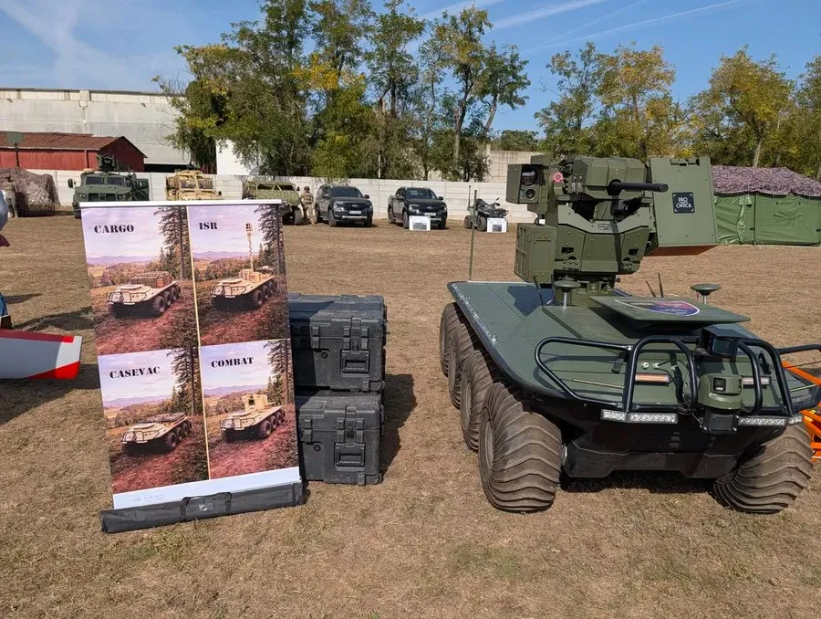 O dronă terestră verde cu roți mari, dotată cu un turn de armament, este poziționată pe un câmp lângă un panou informativ și câteva laduțe negre.