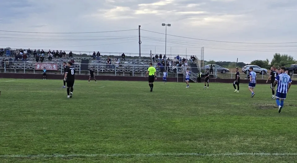 O imagine panoramică a unui teren de fotbal cu jucători în tricouri albastre și negre, un arbitru în galben și spectatori pe tribună.